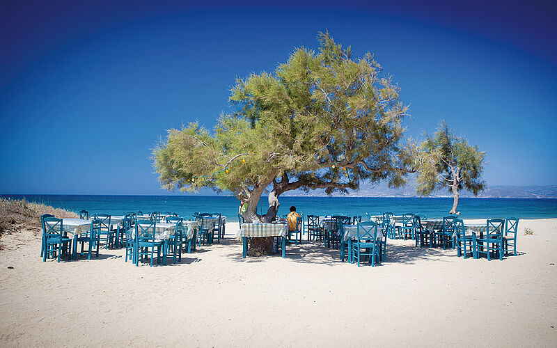 ein Bild mit Blick auf einen Strand auf Naxos mit Tischen, Stühlen und einem Baum